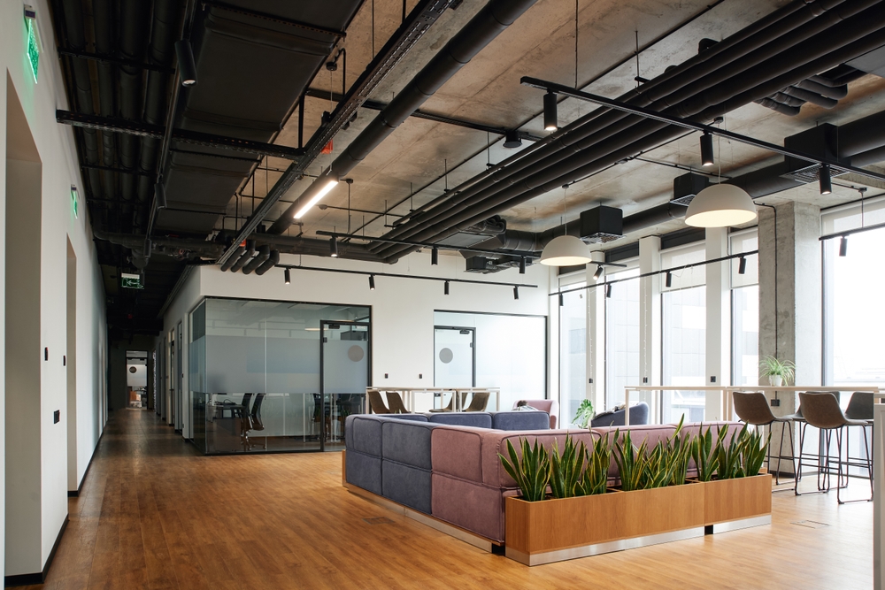 Spacious and contemporary office lounge featuring a soft and comfortable couch, large windows, high chairs, and green plants, designed with climate control systems to ensure optimal comfort and air quality, reflecting a modern workspace organisation concept.